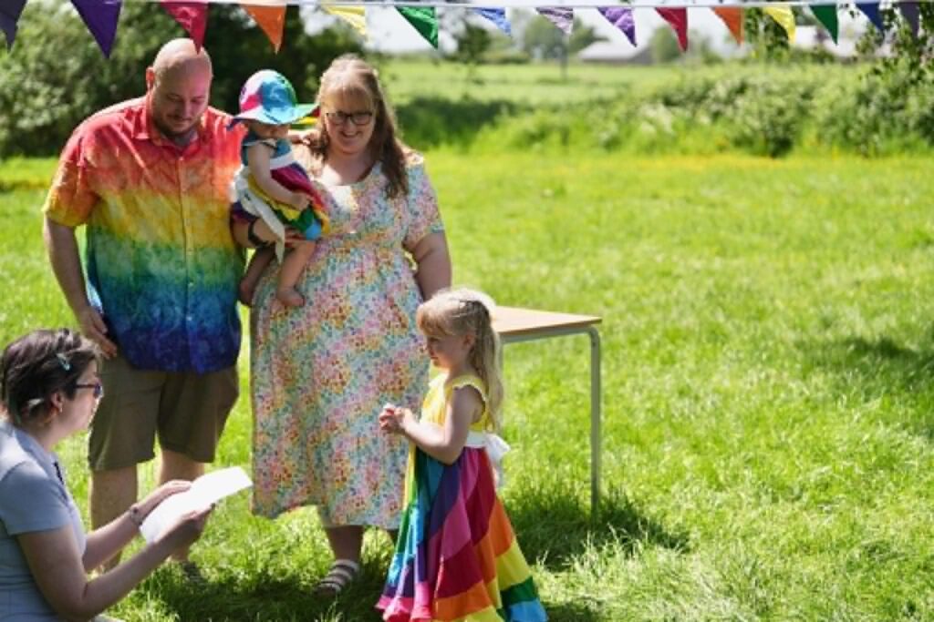 Celebrant leadinga young child through promises for her sister, her parents and sister are standing alongside her in a sunny grassy field with rainbow bunting